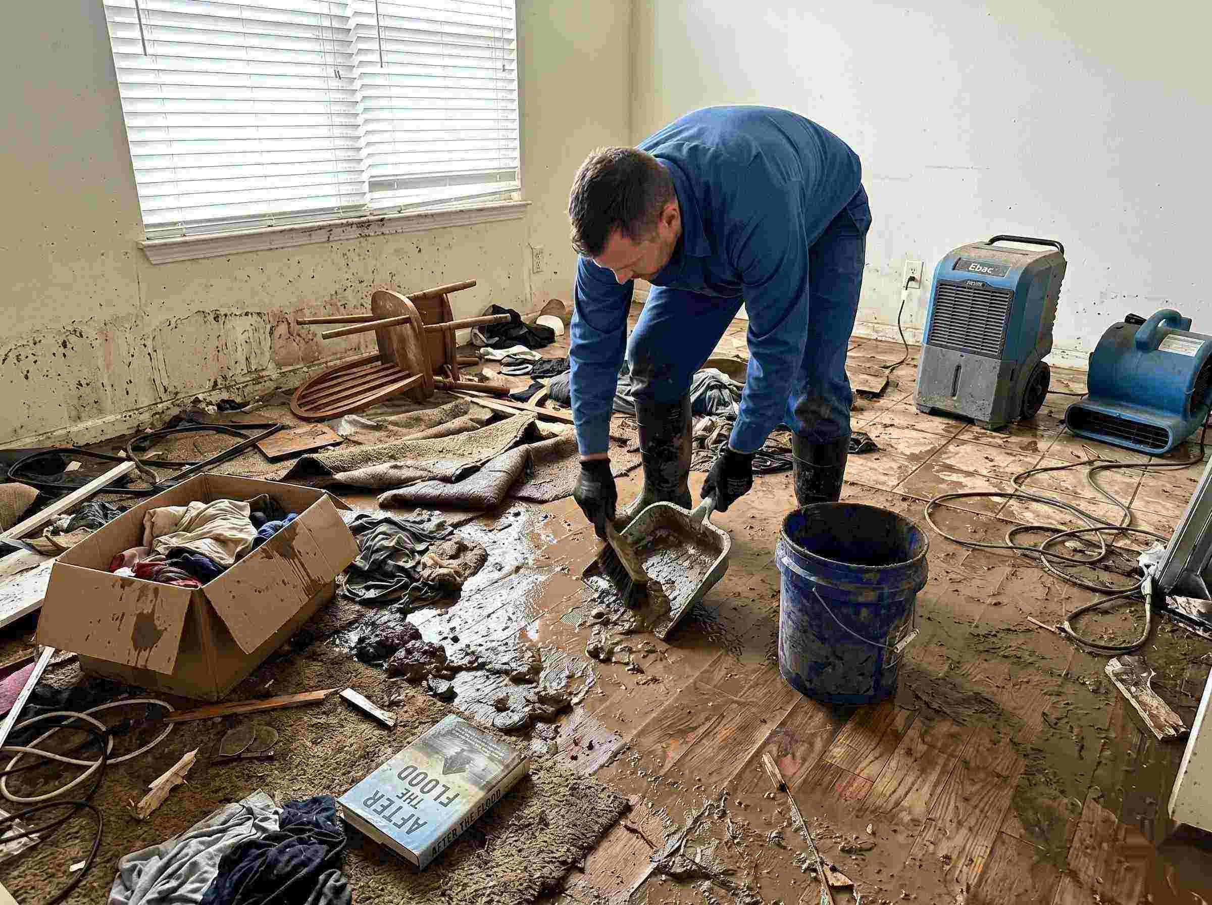 Realistic photo of a technician performing mud and debris removal after a home flood in Baltimore, showing soaked furniture, wet flooring, and scattered debris during post-flood cleanup.